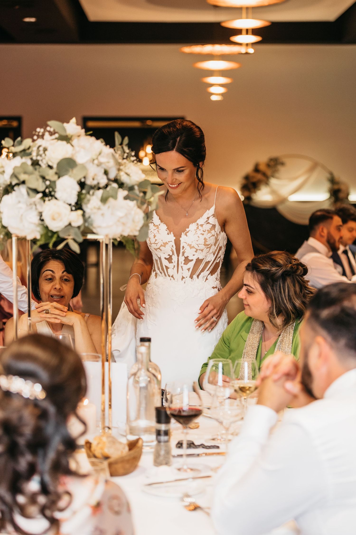 Bride in lace wedding gown smiling and greeting guests at elegantly decorated wedding reception dinner table.