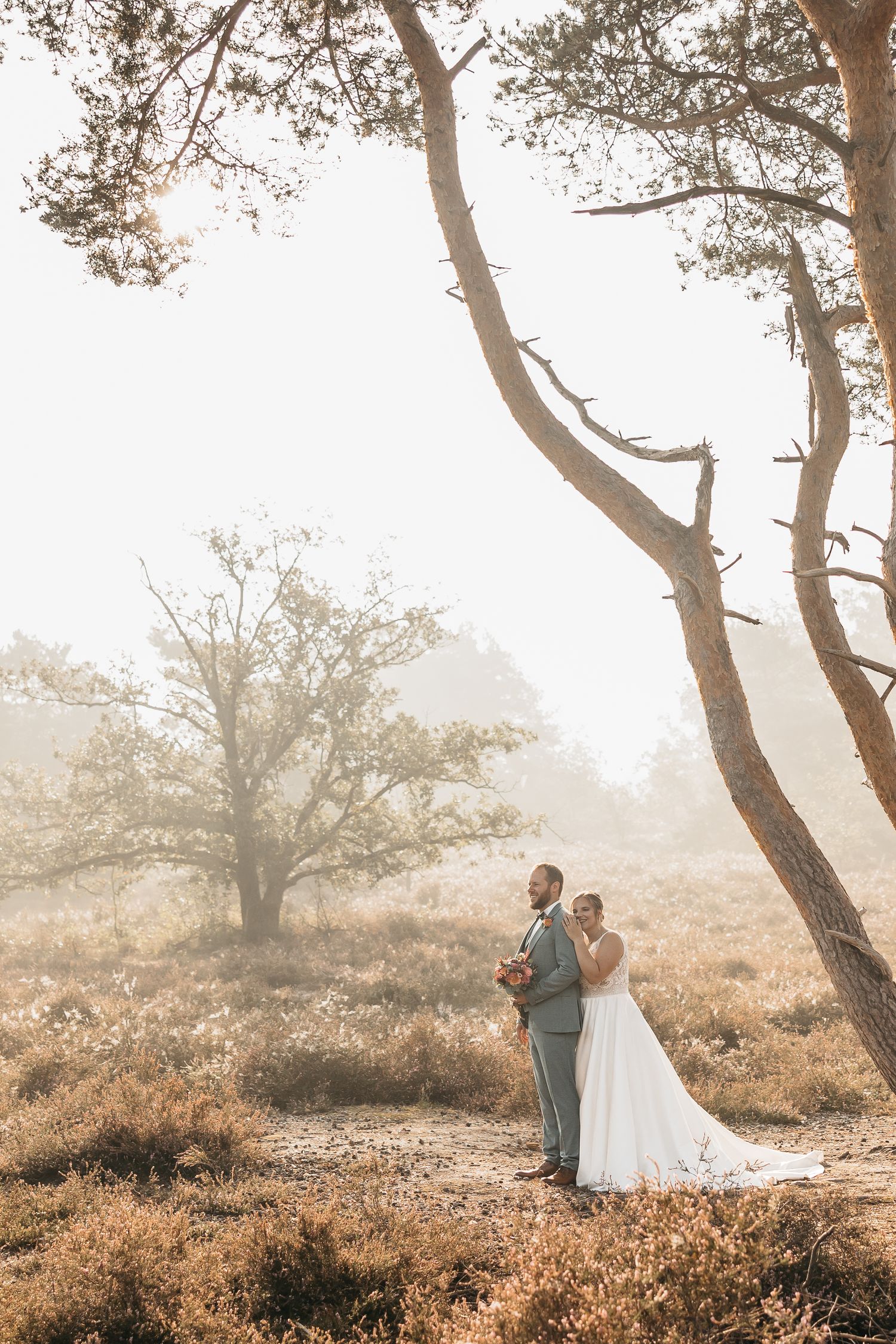 Wedding couple embraces among tall pine trees in a golden misty heathland landscape during their bridal shoot.