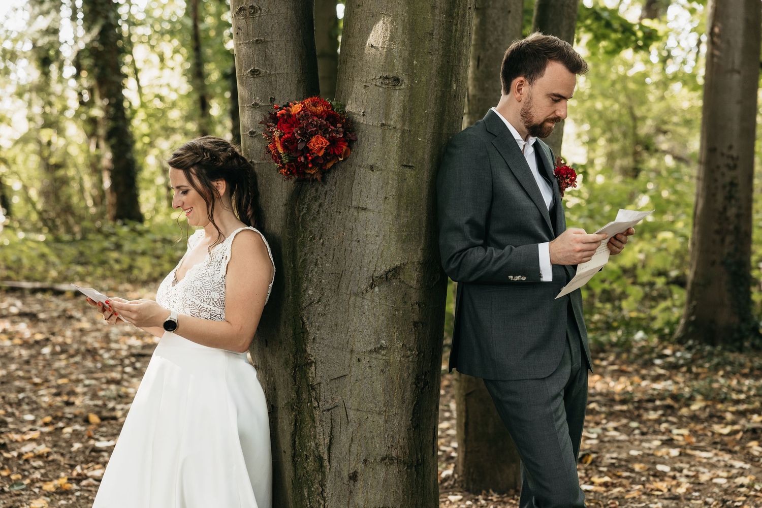 Bride and groom leaning back-to-back against a tree reading their wedding vows privately in a forest.