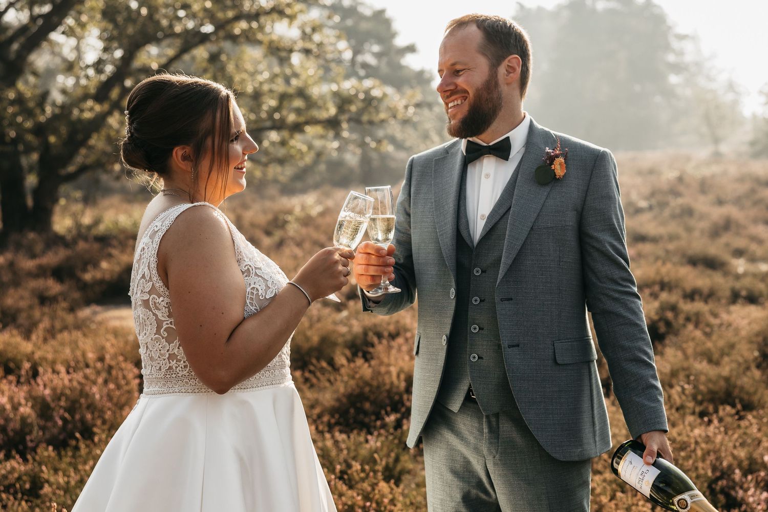 Smiling newlyweds clinking champagne glasses in a sun-drenched heather field after their wedding.