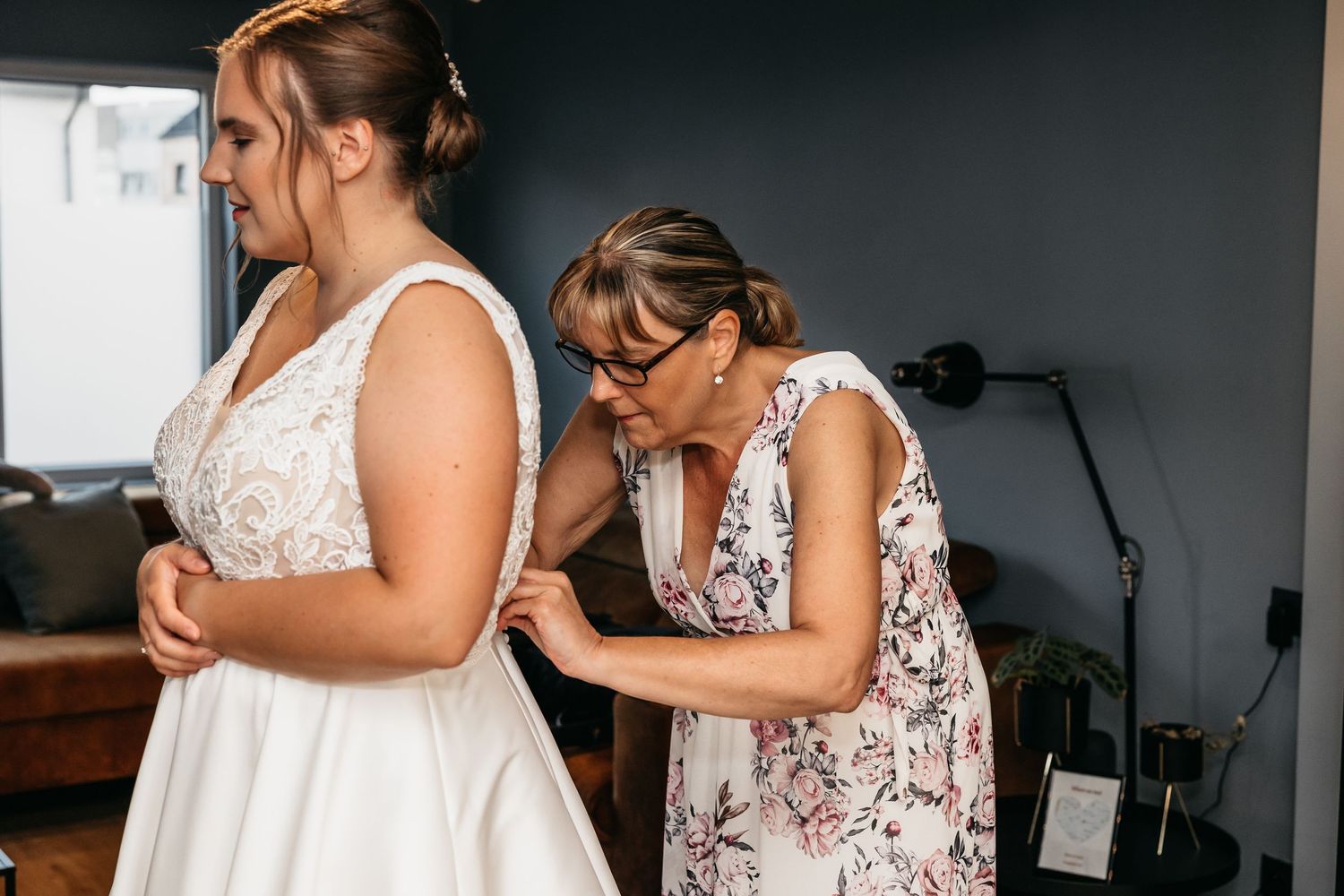 Woman in floral dress helping bride fasten the back of her white lace wedding gown indoors.