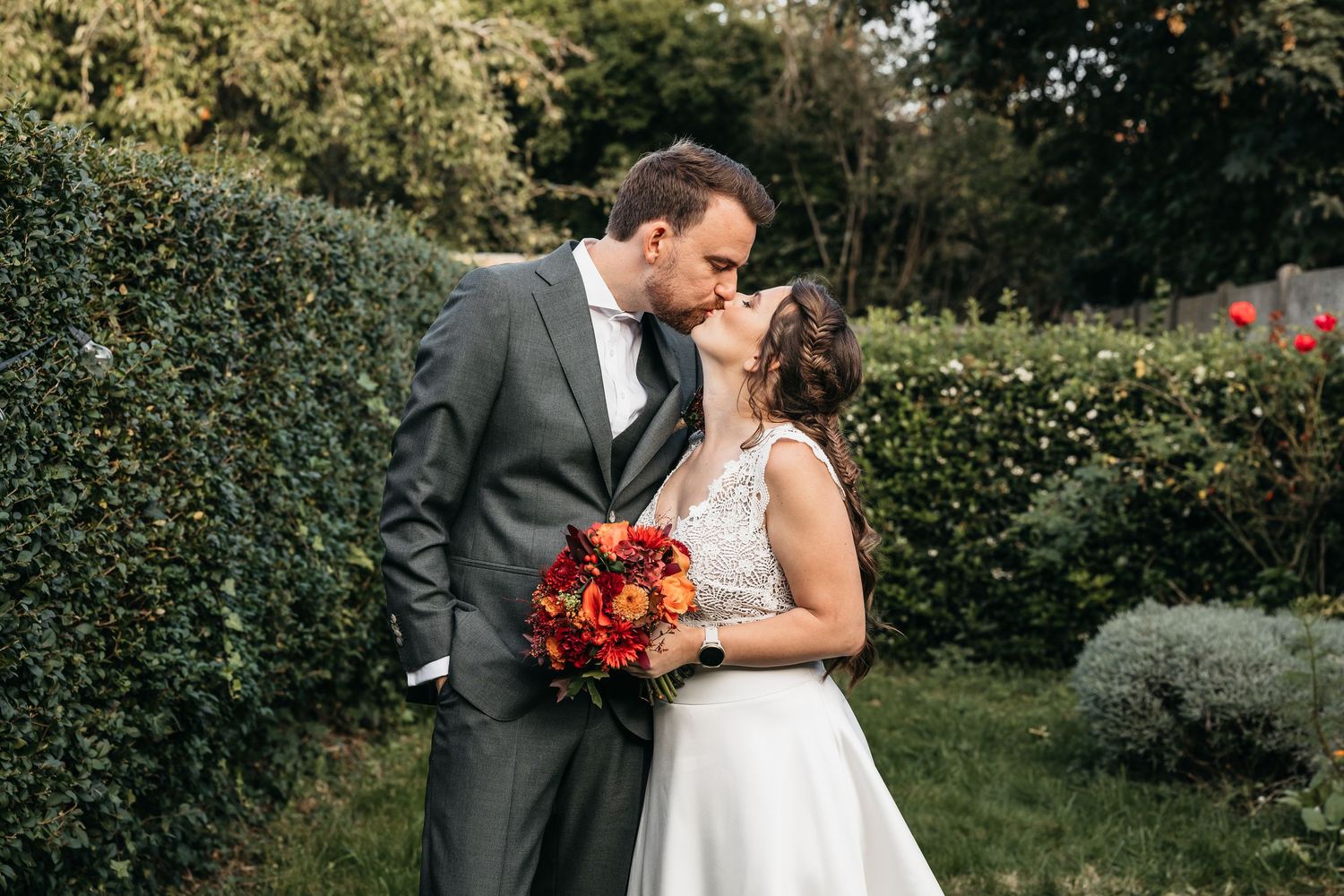 Newlyweds sharing a romantic kiss outdoors, bride holding vibrant red and orange bouquet.