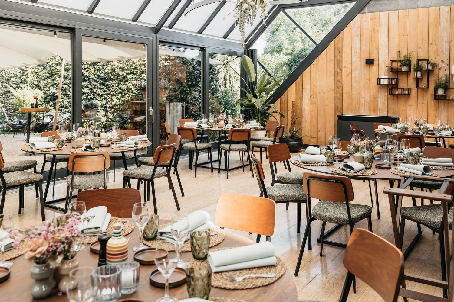 Bright greenhouse restaurant with round tables set for a wedding reception, featuring wooden walls and lush greenery.