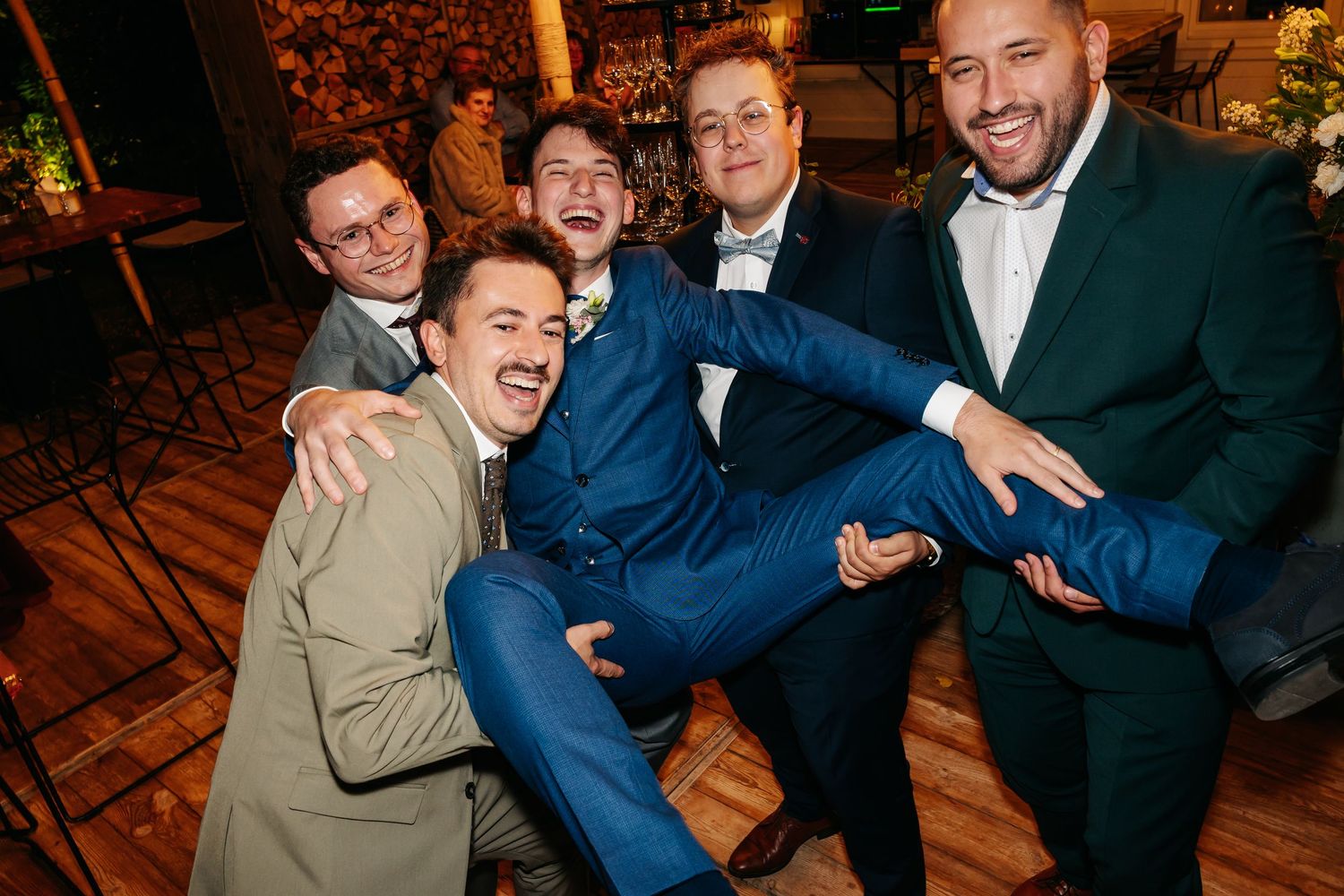 Five men in suits laughing and posing playfully together at a festive wedding reception.