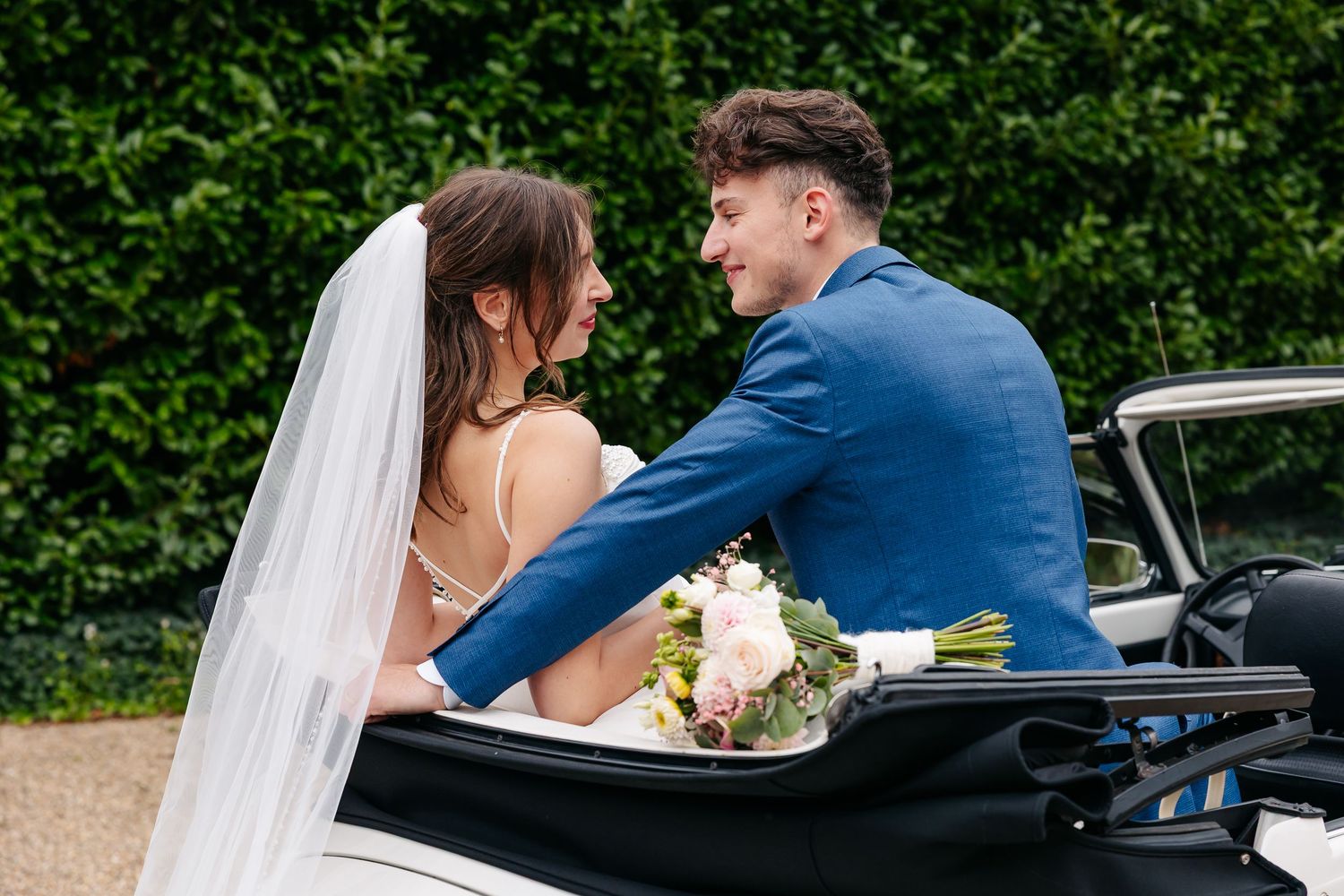 Bride and groom share an intimate moment seated together in classic white convertible wedding car.