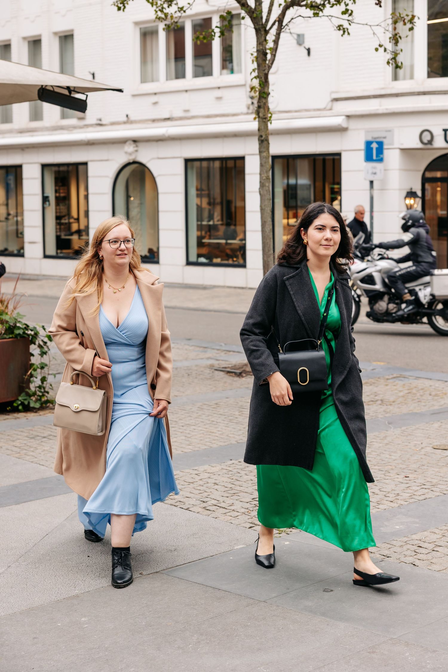Two women walking city street, one in blue satin dress with camel coat, other in green dress with black blazer.