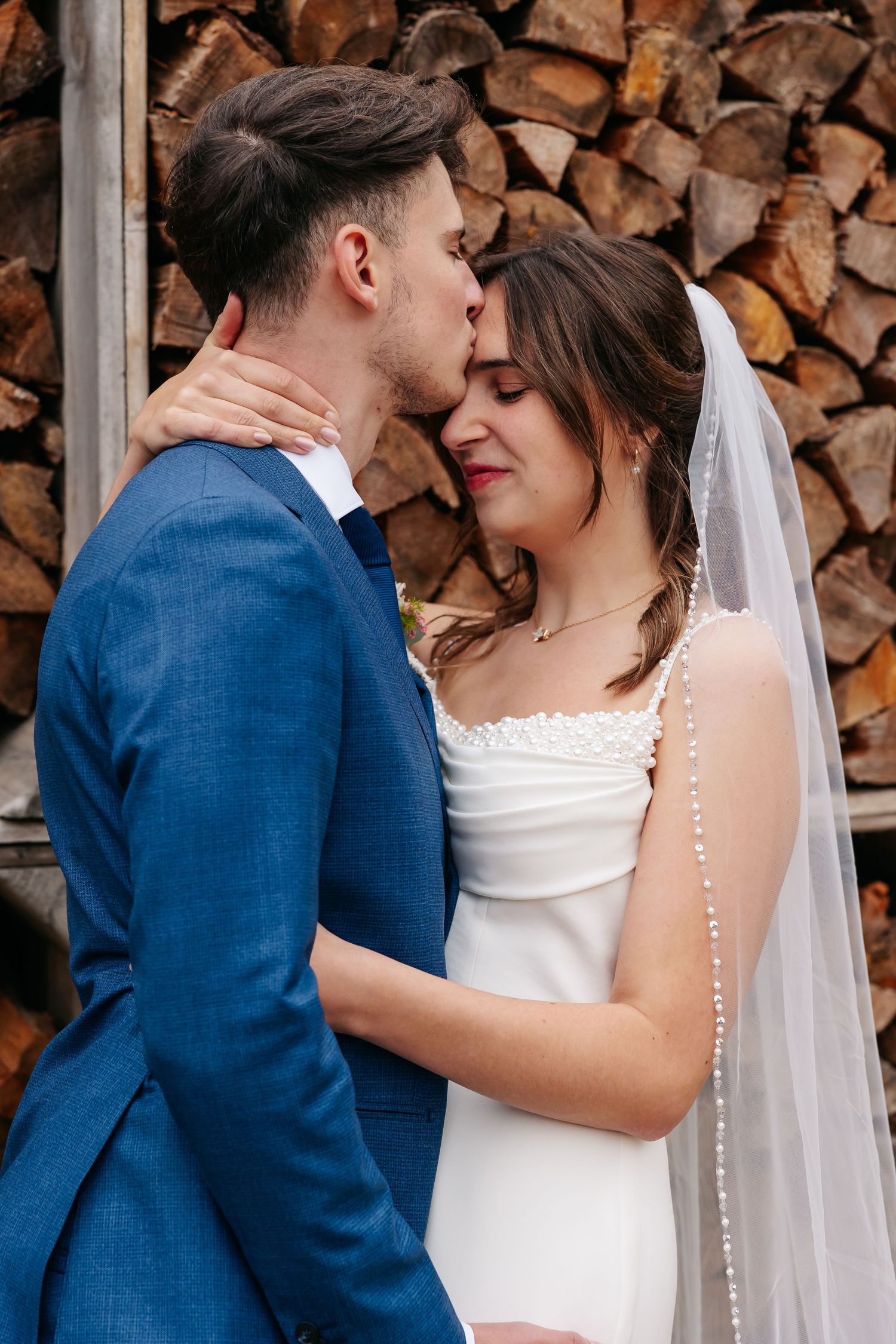 Groom in blue suit kissing bride in white dress and veil against rustic wood background.