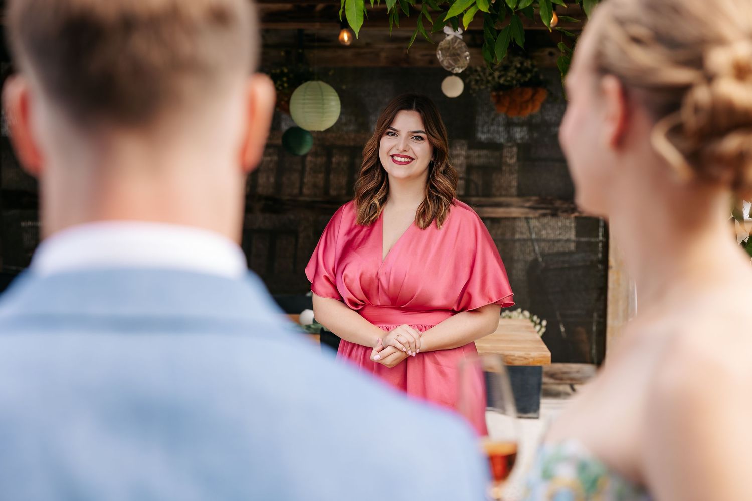 Woman in pink dress smiling at wedding guests during outdoor reception.