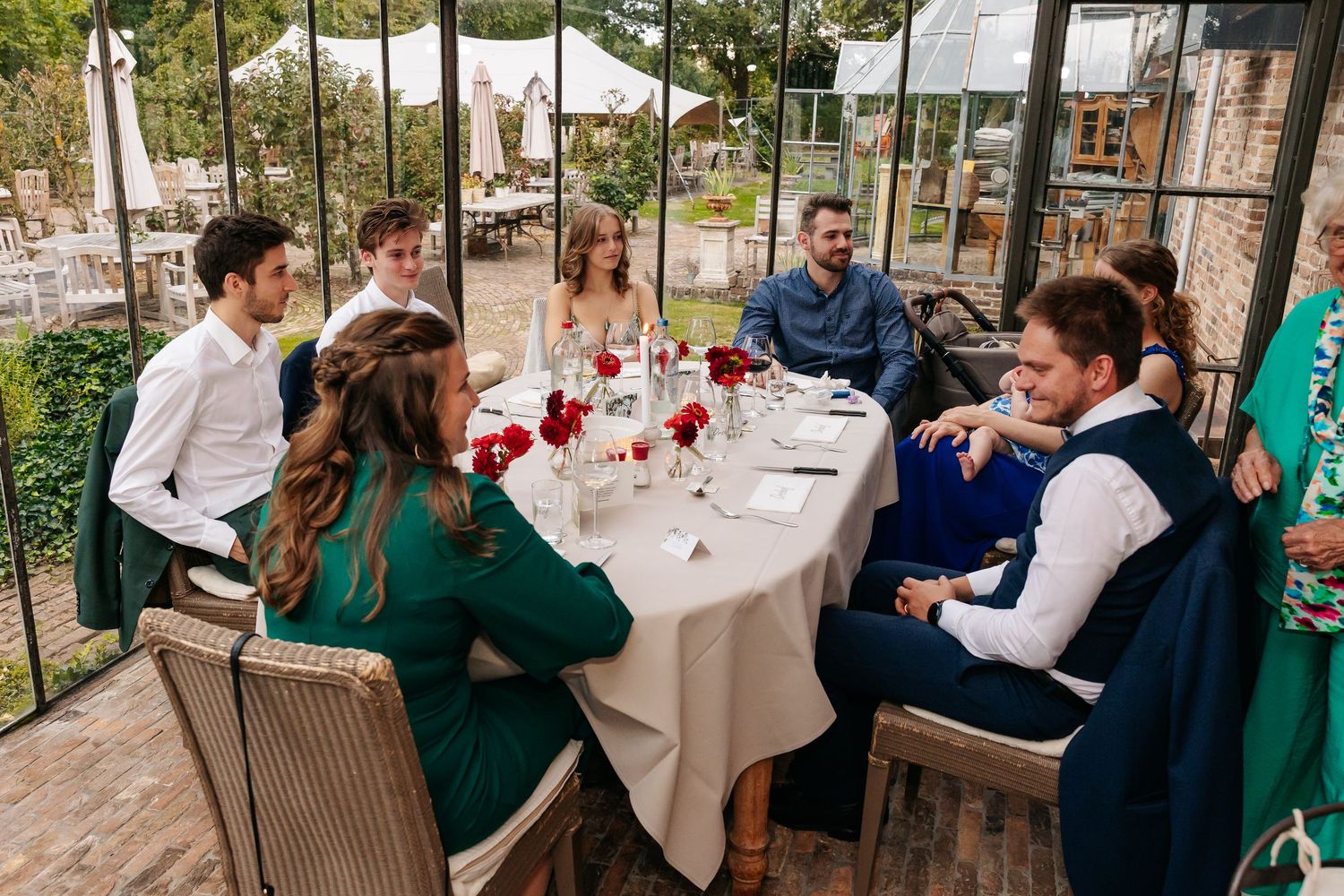 Small group of elegantly dressed wedding guests conversing at a decorated table in a glass conservatory.