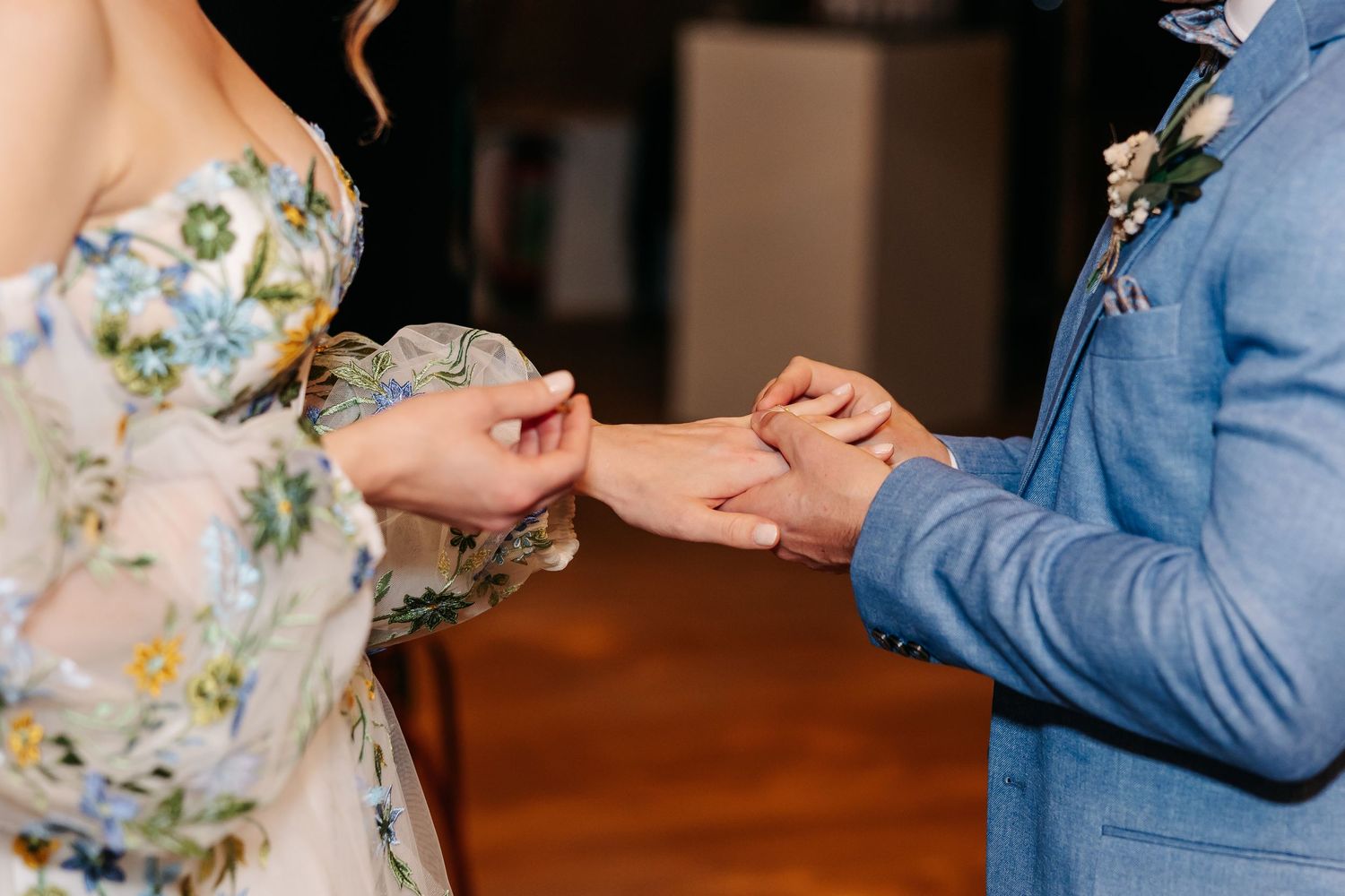 Close-up of couple exchanging wedding rings during marriage ceremony, floral dress detail visible.