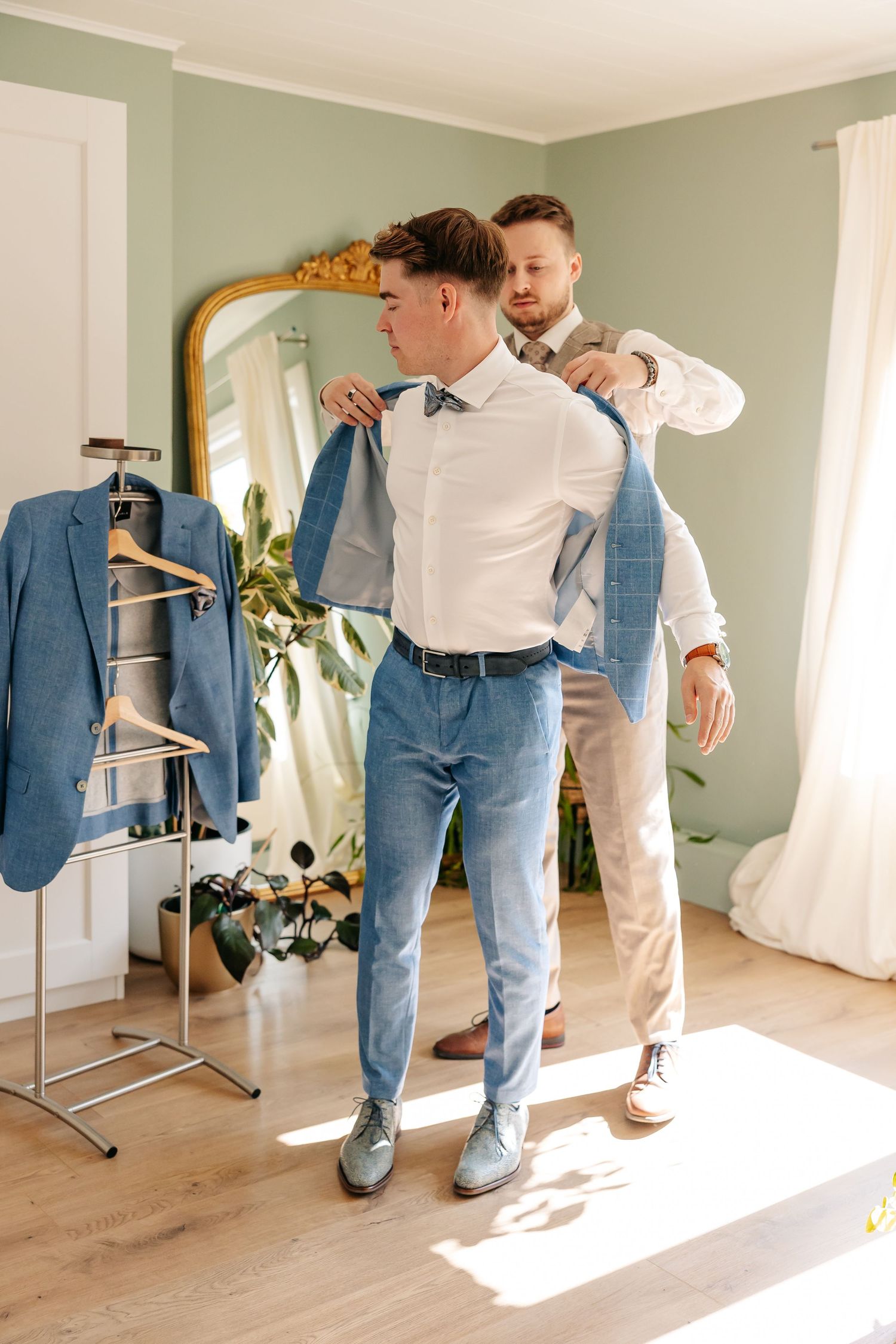 Groomsman helping groom put on light blue suit jacket near gold mirror.