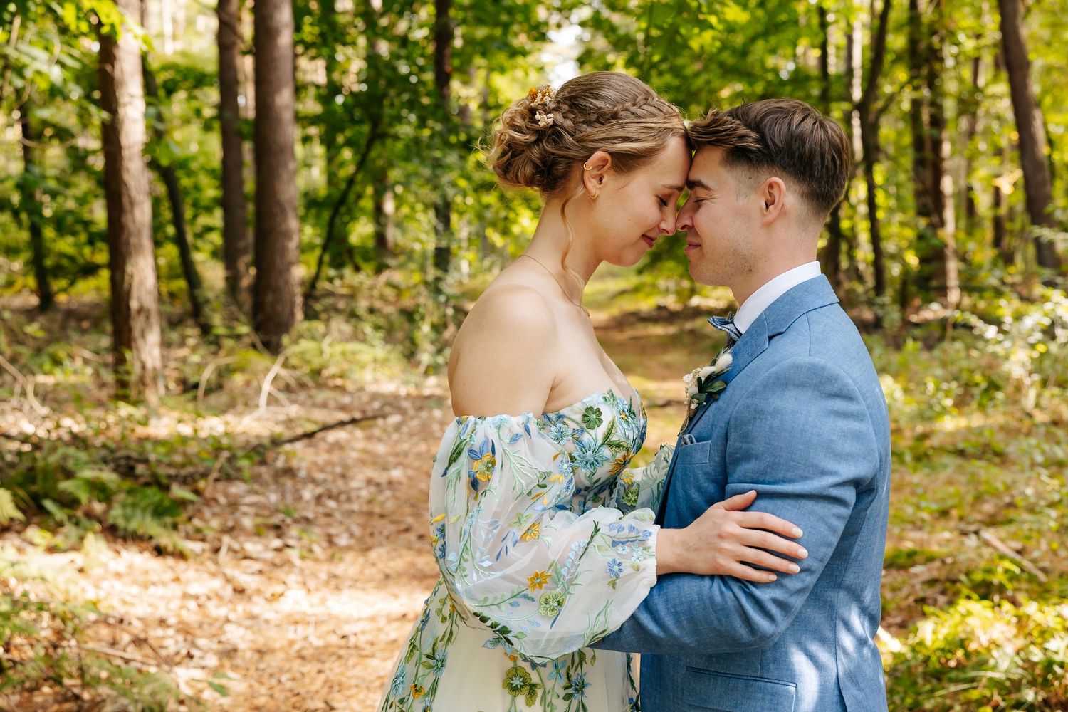 Bride and groom touching foreheads tenderly on sunny forest path.
