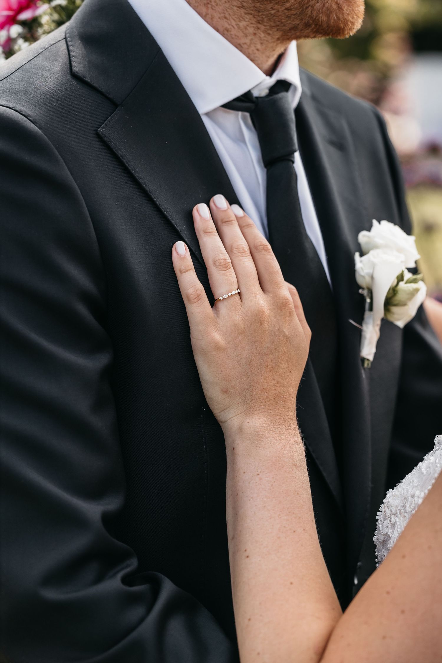 Close-up of brides hand with gold engagement ring resting on grooms black suit jacket lapel.