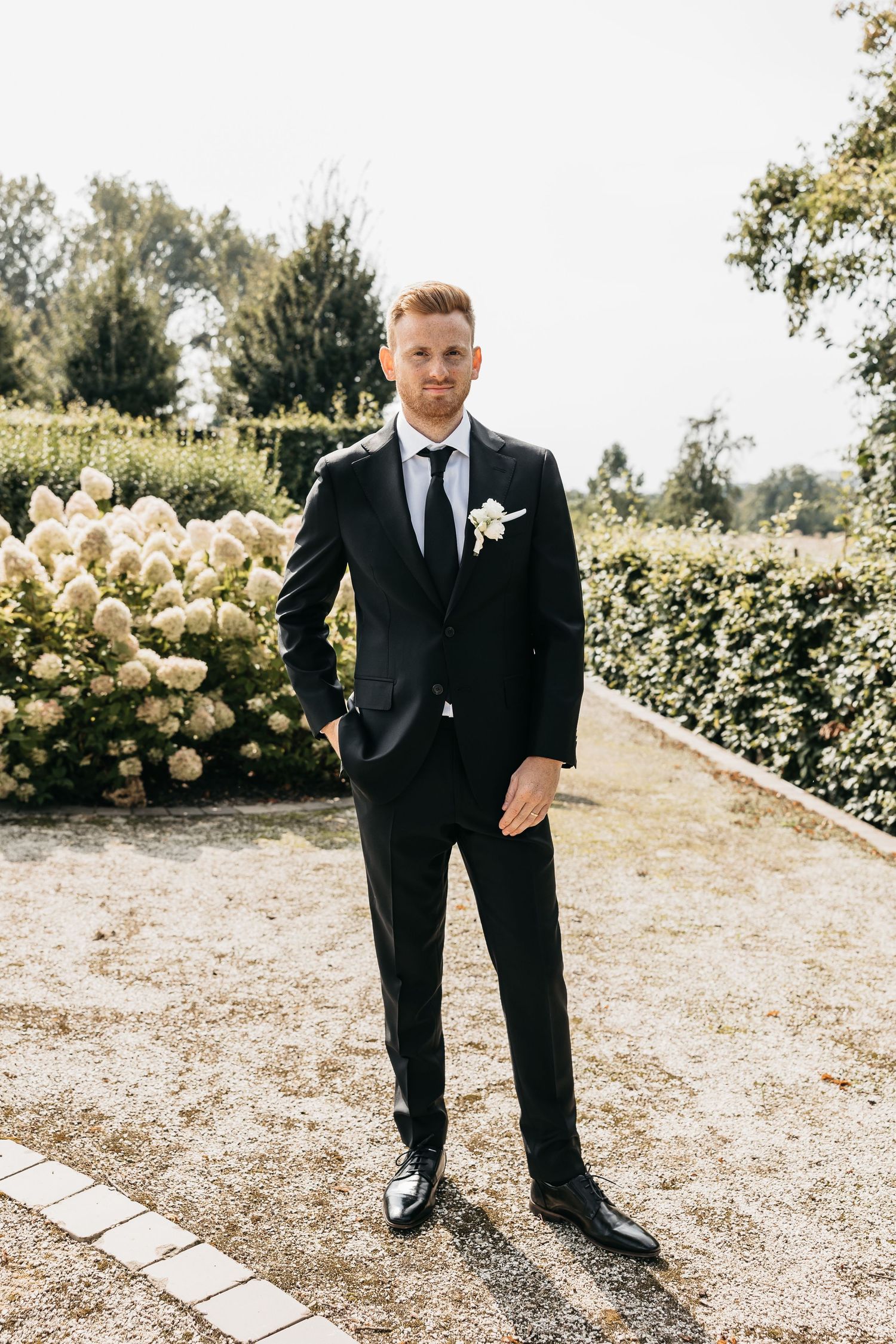 Groom in black suit with white boutonniere standing confidently on a gravel garden path.