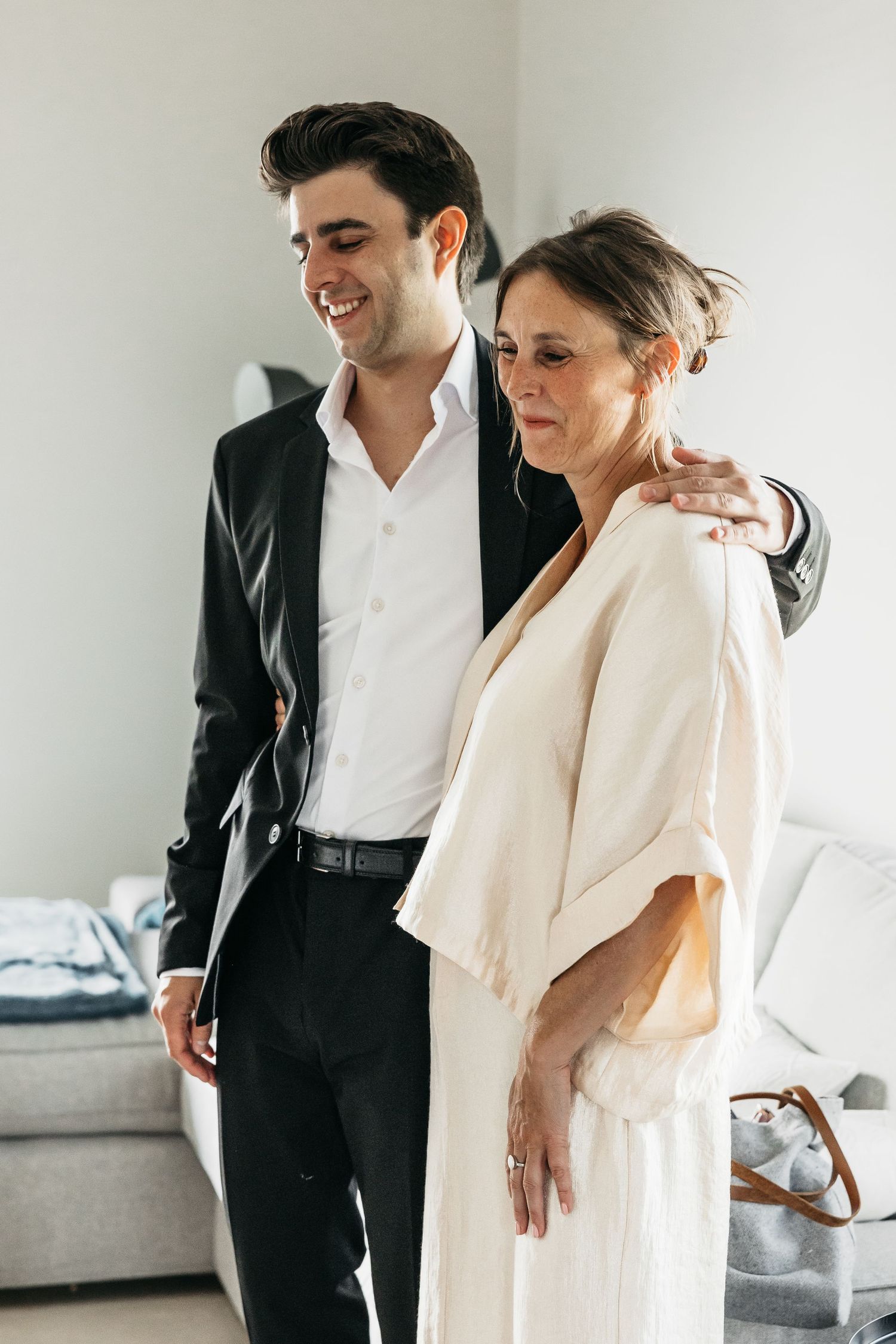 Smiling young man in black suit embracing an older woman in cream dress during wedding preparations.