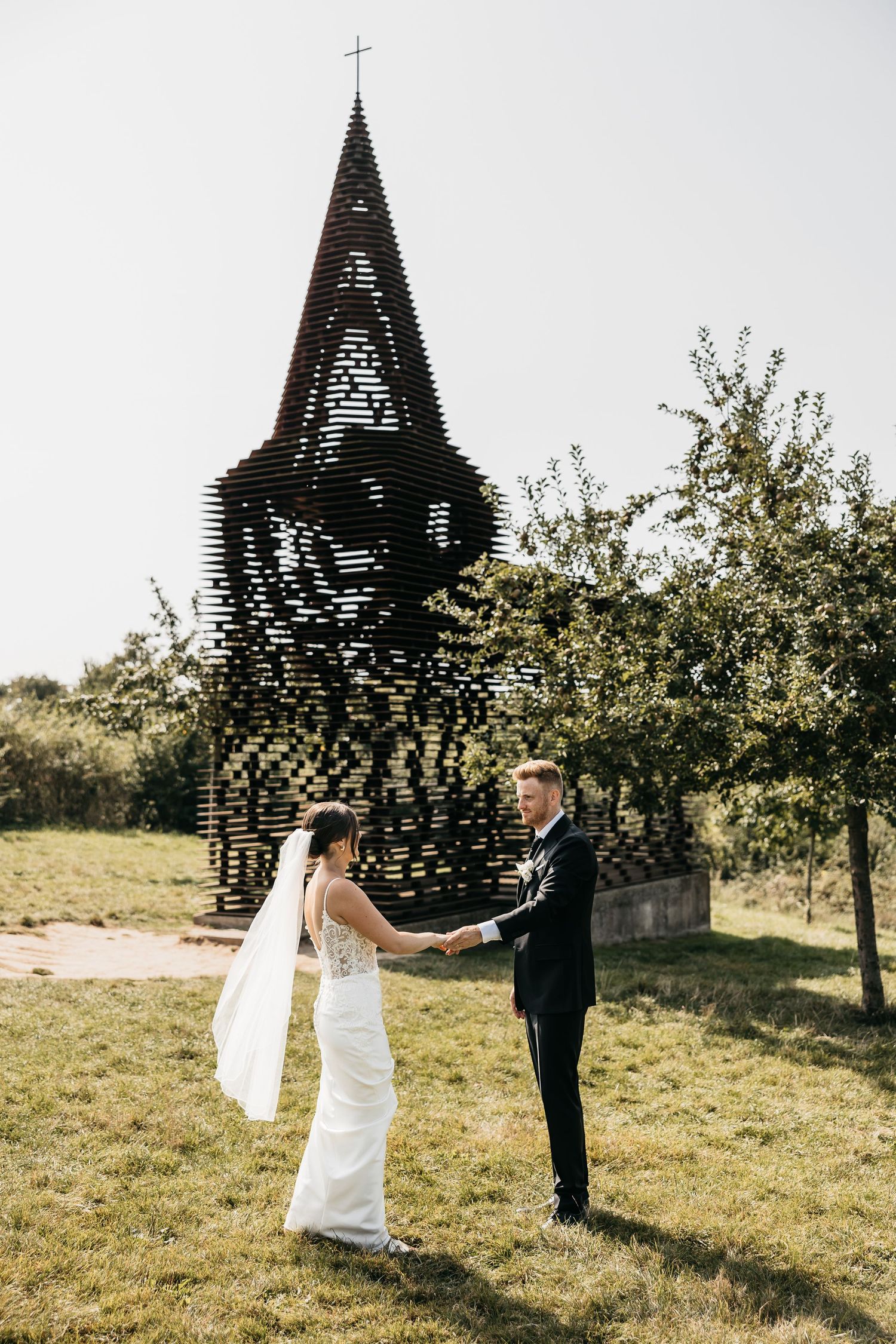 Bride and groom holding hands facing each other near a striking geometric church tower sculpture.