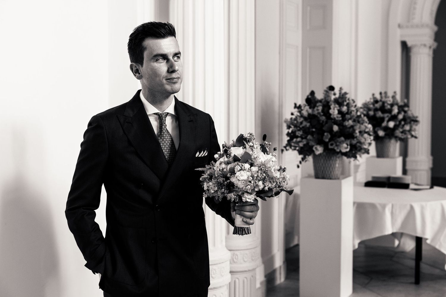 Black and white photo of groom holding floral bouquet standing in classic wedding venue.