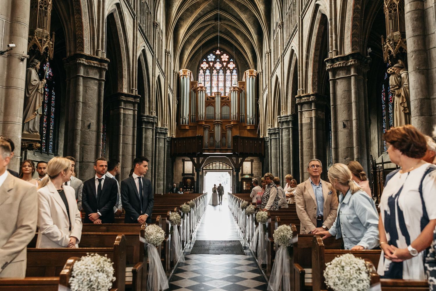 Wide shot of bride walking down ornate Gothic cathedral aisle lined with guests and white floral decorations.