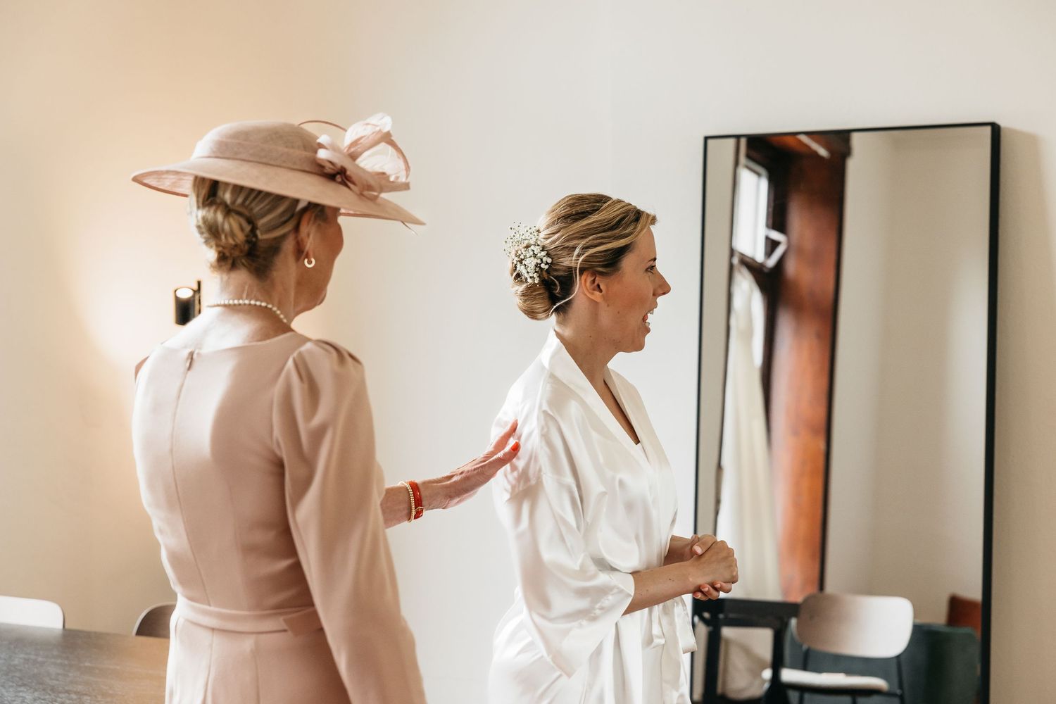 Woman in pink hat and dress assisting bride in white robe near a mirror on wedding day.