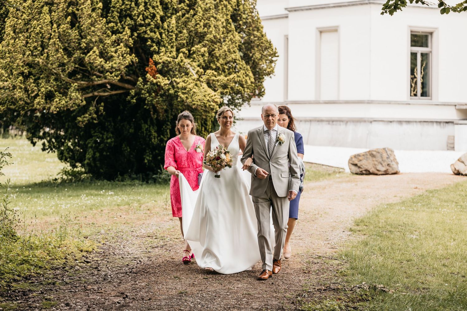 Bride in white dress walks with two guests along garden path beside a grand white chateau wedding venue.