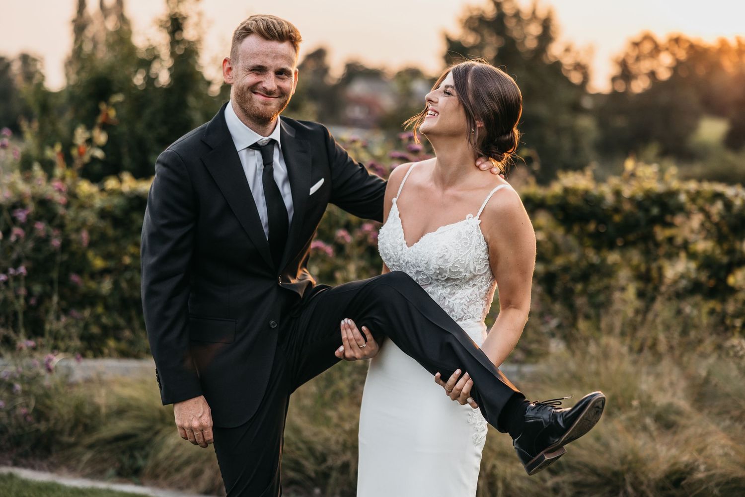 Laughing bride and groom playfully pose together outdoors, groom lifting bride's leg in a joyful sunset moment.