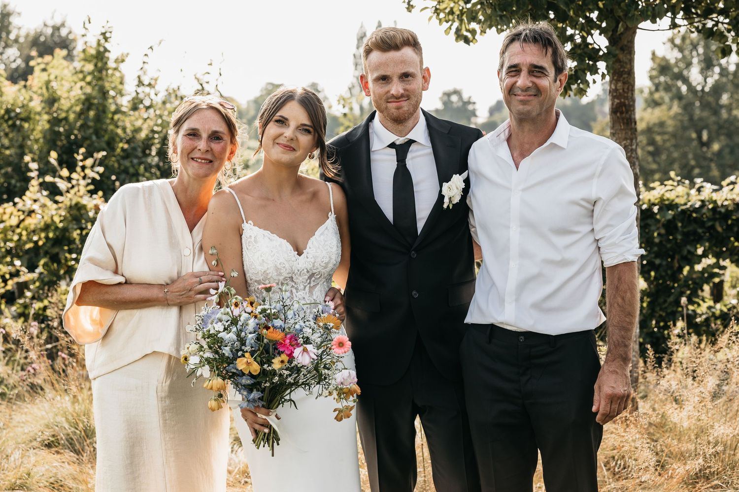Bride in white lace gown with wildflower bouquet poses with groom and two family members in sunny outdoor setting.
