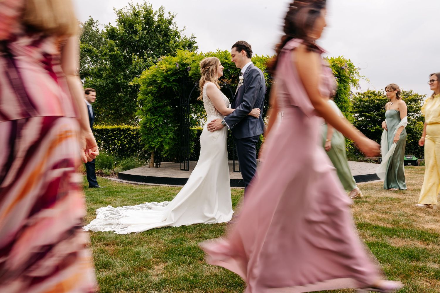 Bride and groom share a romantic moment as wedding guests walk past in colorful dresses outdoors.