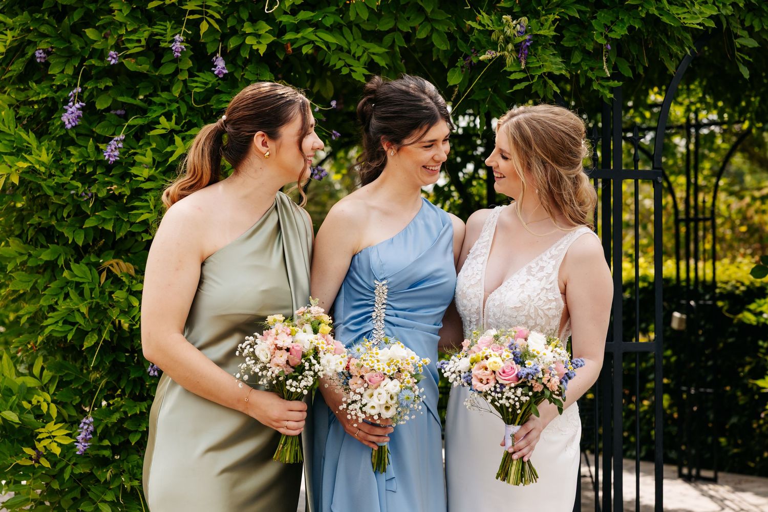 Three women in wedding party laughing together, wearing sage, blue, and white dresses with bouquets.