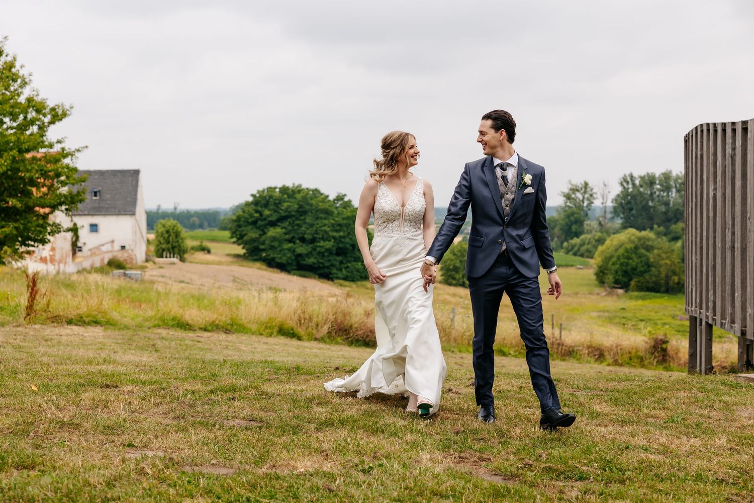 Happy bride and groom holding hands and smiling at each other while walking across a lush green field.