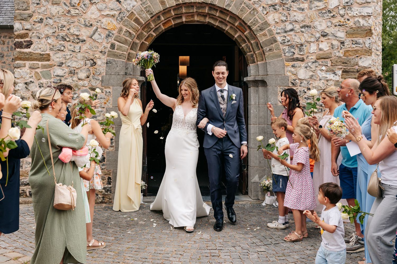 Bride raises bouquet as couple exits rustic stone church surrounded by cheering wedding guests.