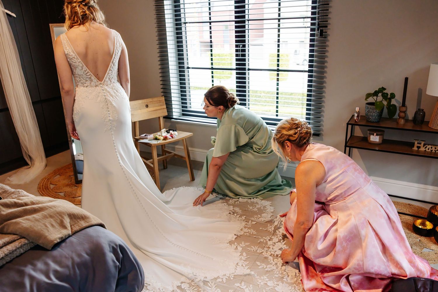 Two bridesmaids in sage and pink dresses carefully arranging the lace train of a bridal gown by a window.
