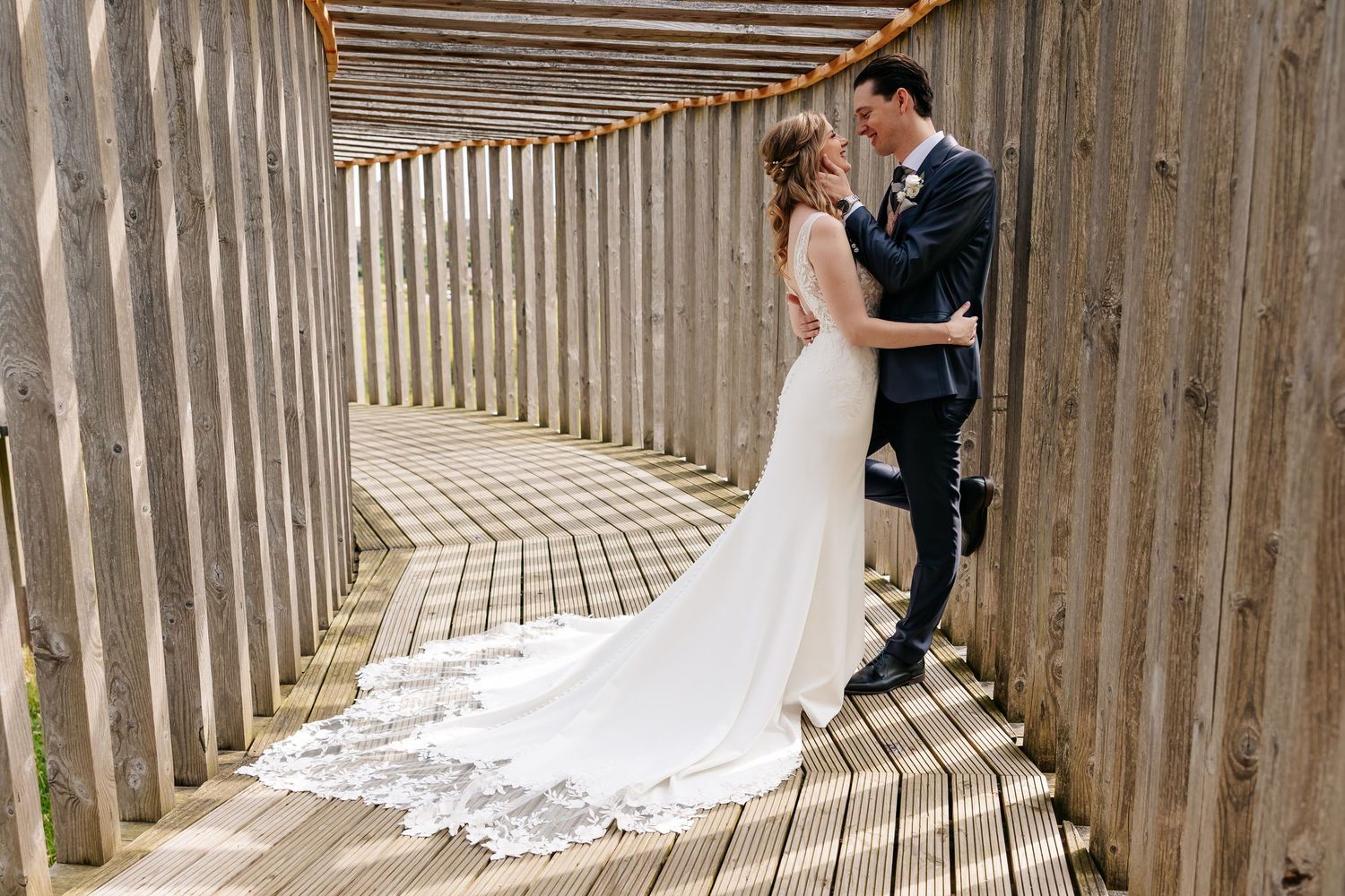 Bride and groom embrace romantically in sunlit wooden pergola walkway on wedding day.
