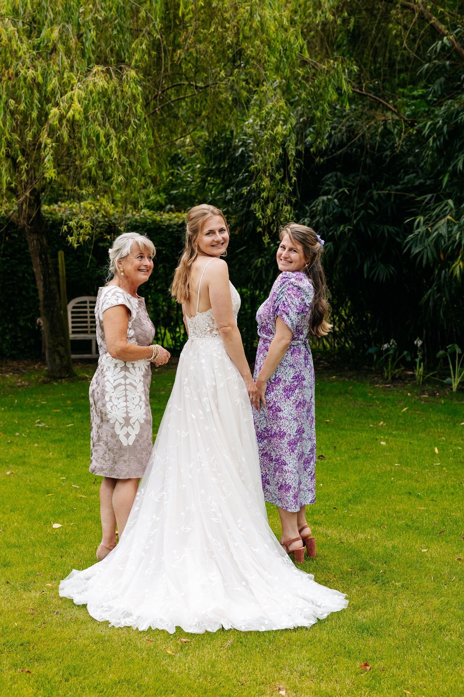 Bride in backless lace wedding gown posing with two women on green lawn under willow tree.