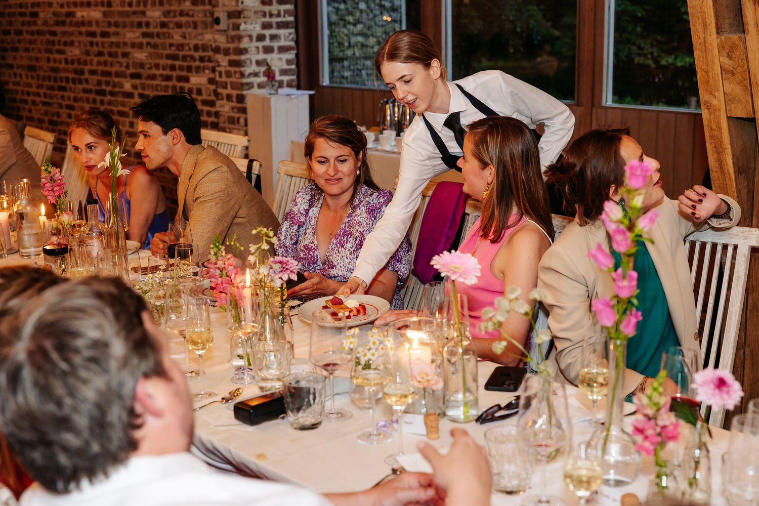 Waitress serves food to smiling wedding guests at elegantly decorated candlelit dinner reception table.
