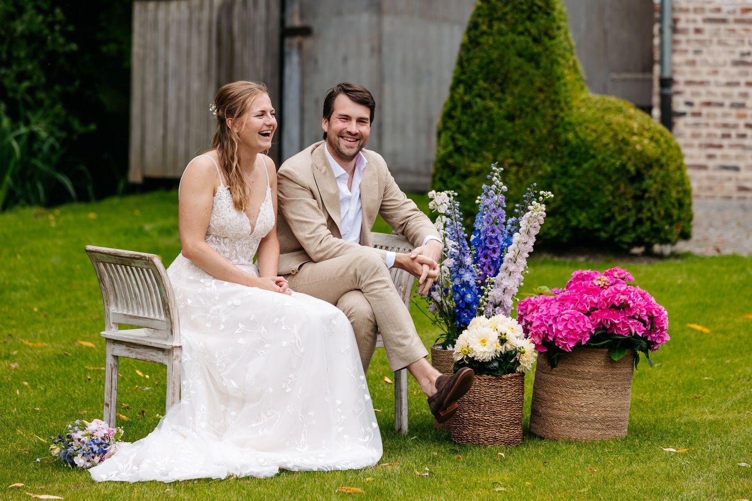 Bride and groom laughing together on a bench surrounded by colorful flowers in a garden.