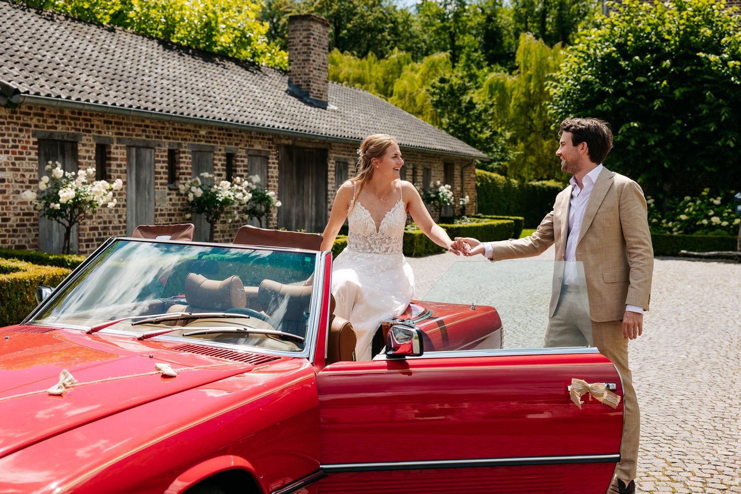 Bride sitting on red vintage Mercedes convertible holding grooms hand at sunny outdoor wedding.