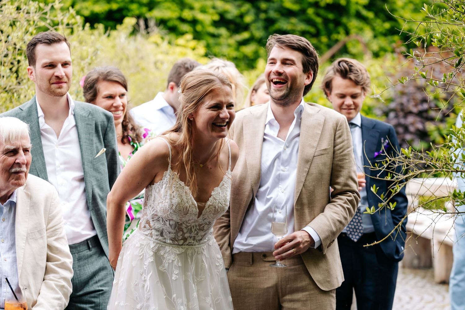 Laughing bride in lace dress and groom in beige suit walk with wedding guests in sunny garden.