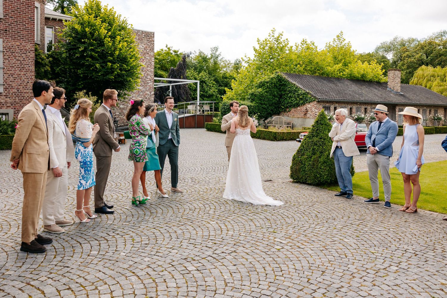 Wedding couple greeting guests on a cobblestone courtyard surrounded by lush green trees and hedges.