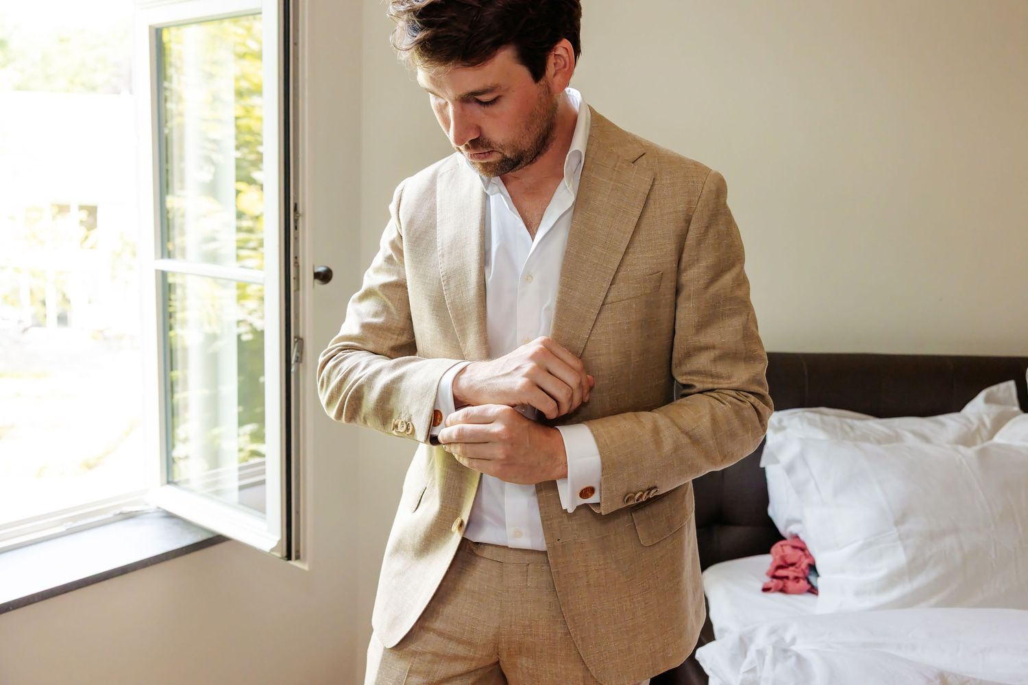 Handsome groom in beige linen suit adjusting cufflinks in bright bedroom before wedding ceremony.