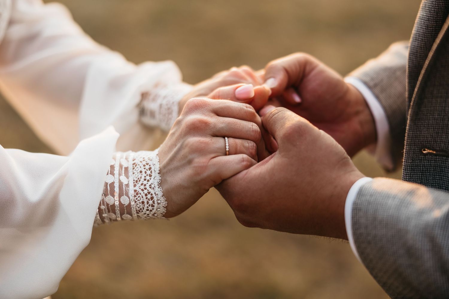 Close-up of bride and groom holding hands, wedding ring visible on brides finger during outdoor ceremony.