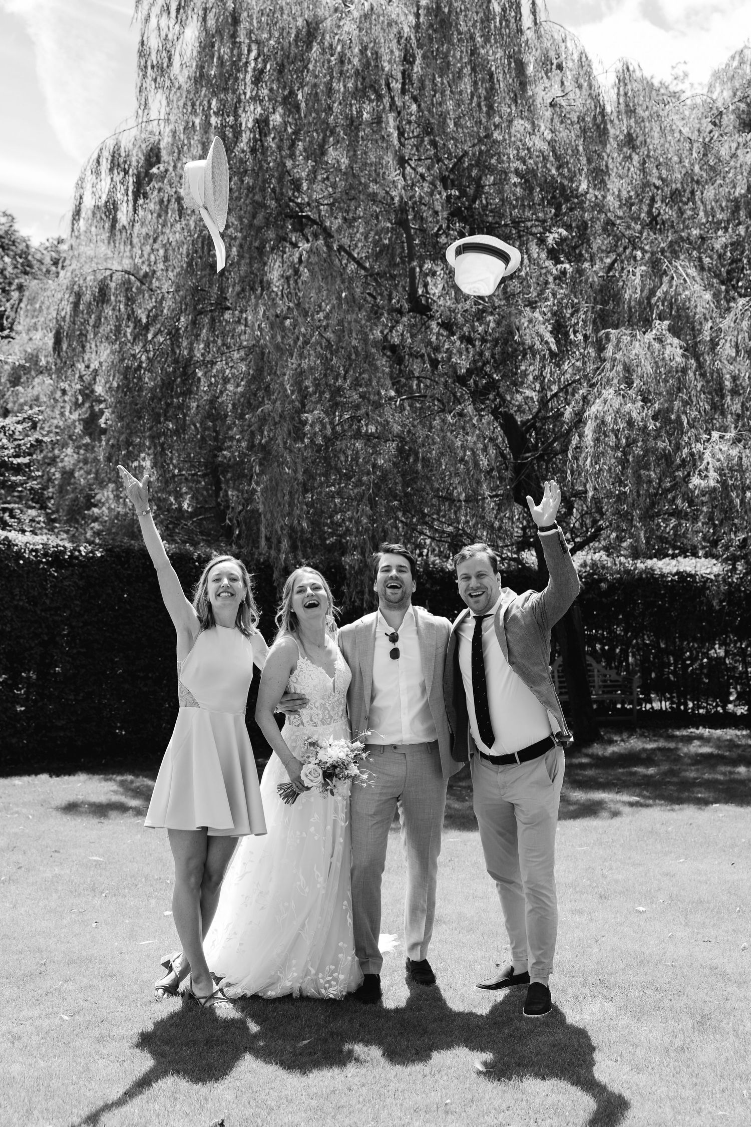 Black and white photo of a joyful wedding party tossing hats in the air outdoors.