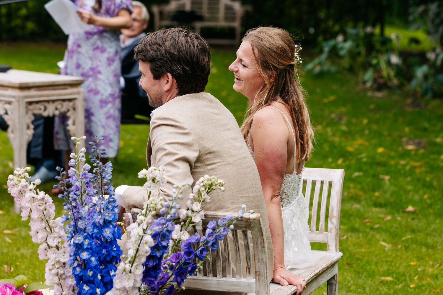 Bride and groom sit together at outdoor wedding ceremony surrounded by colorful blue and purple flowers.