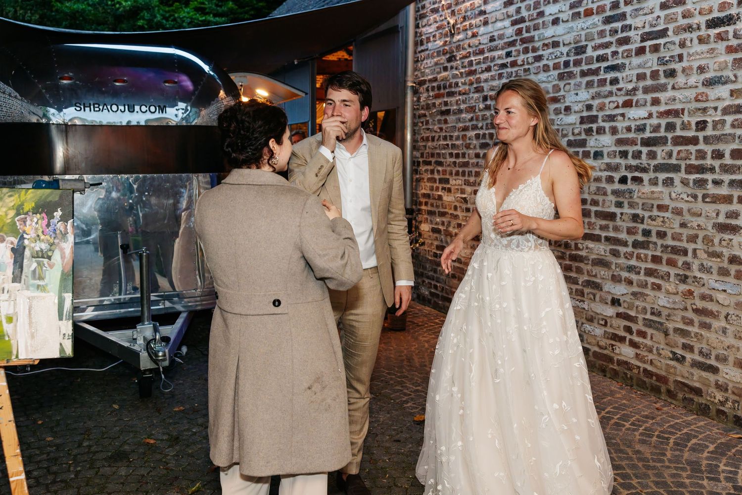 Bride in white gown chatting with guests near brick wall at elegant wedding reception.