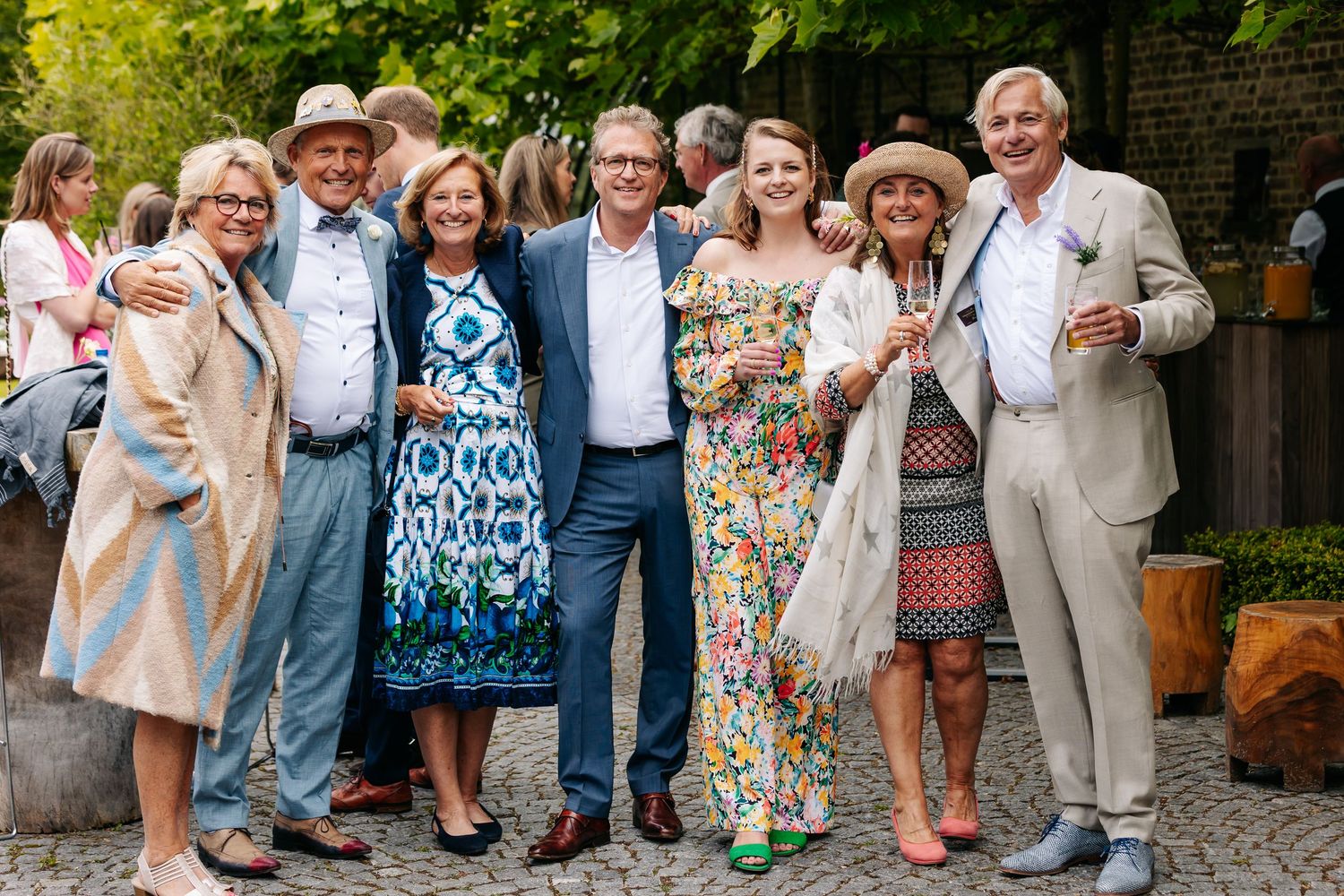 Seven elegantly dressed wedding guests posing together and smiling in a garden.
