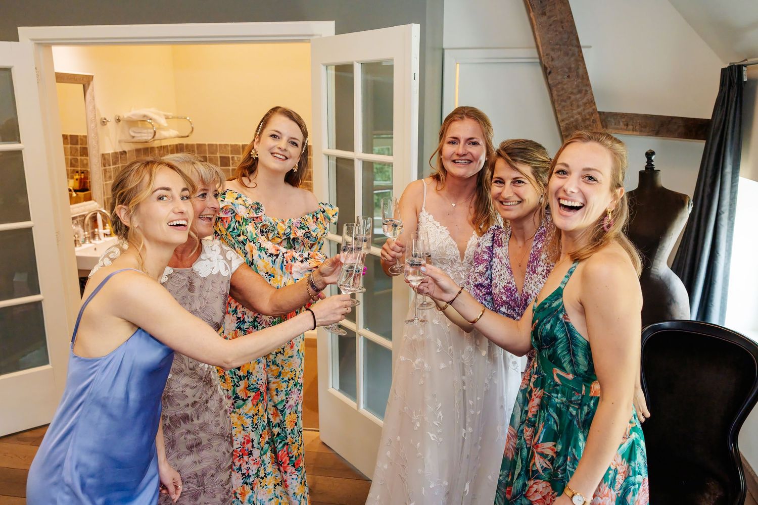 Group of five women in colorful dresses toasting with champagne glasses indoors at a celebration.
