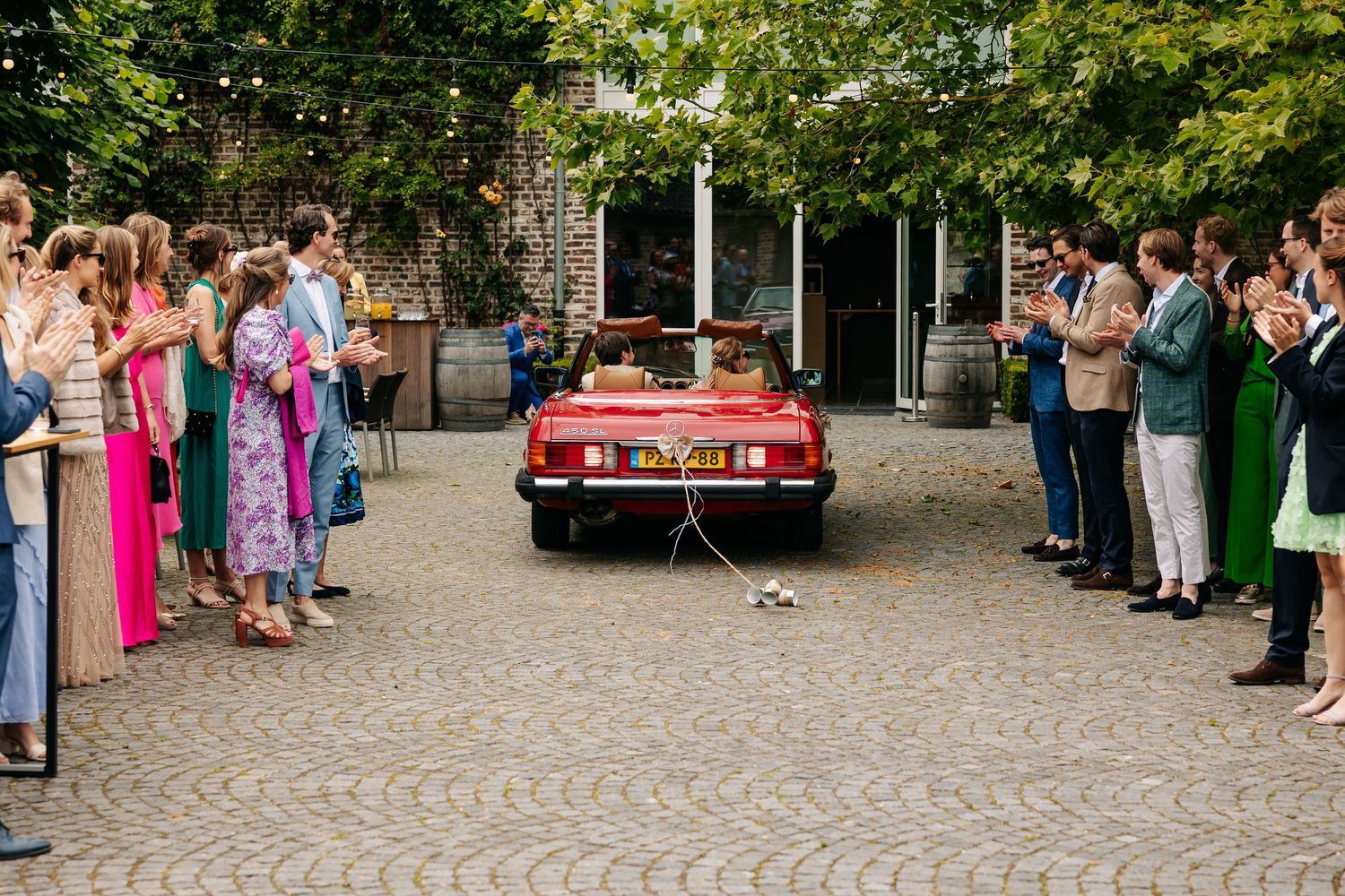 Newlyweds departing in red vintage convertible as wedding guests applaud along cobblestone courtyard of rustic venue.