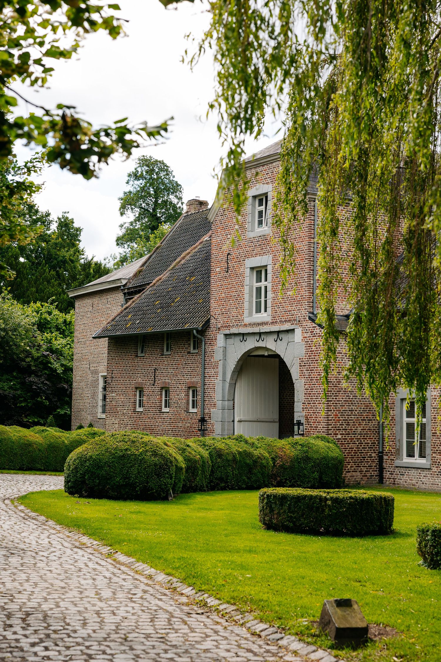 Historic brick manor house with arched gateway, manicured hedges, cobblestone driveway, and weeping willow tree on a cloudy day.