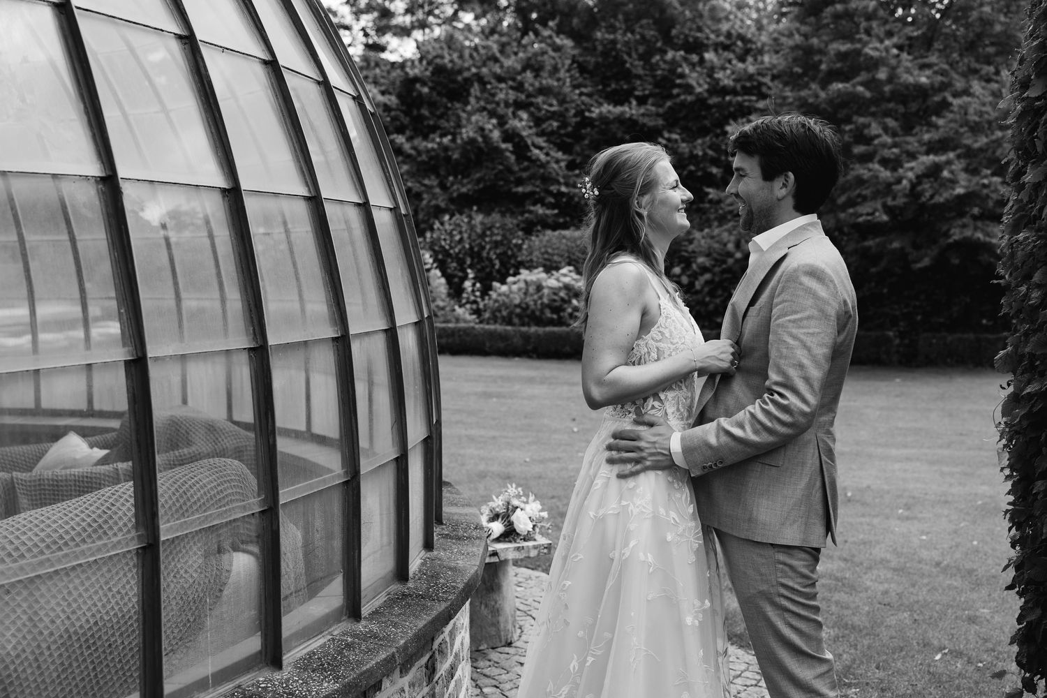 Black and white photo of bride and groom embracing near a greenhouse on their wedding day.