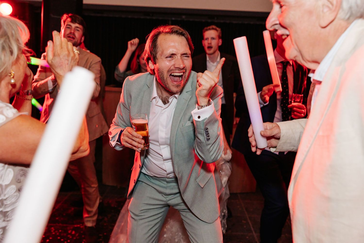 Excited man in mint green suit celebrates energetically with glow sticks at a vibrant wedding party.