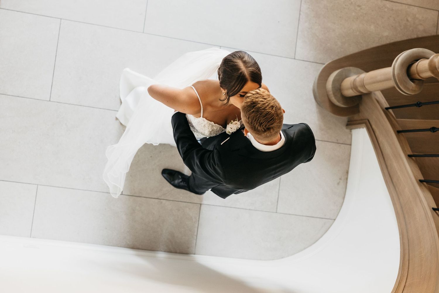 Overhead view of bride and groom embracing at bottom of elegant curved staircase.